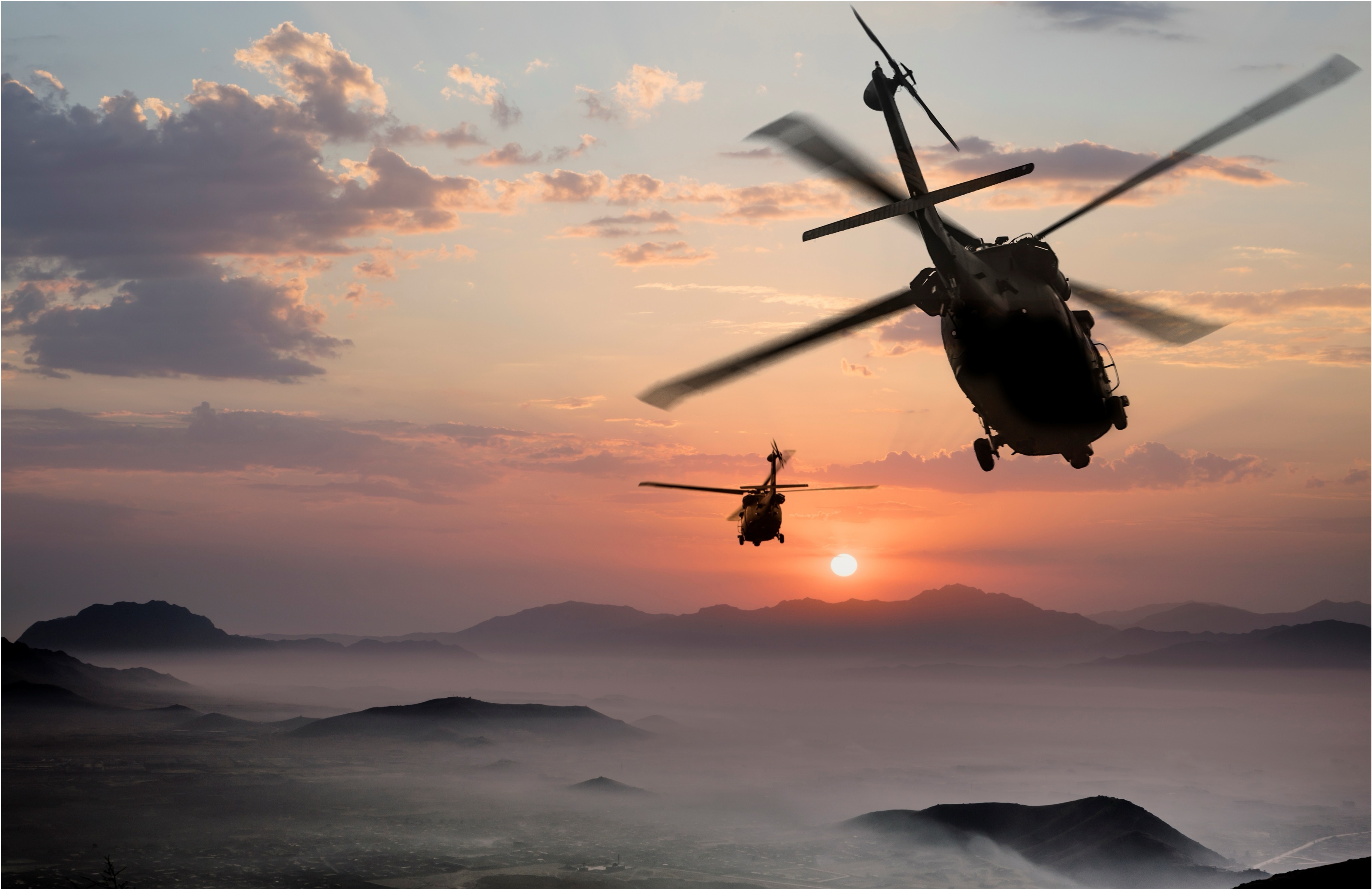 Two military helicopters flying over mountainous terrain at sunset